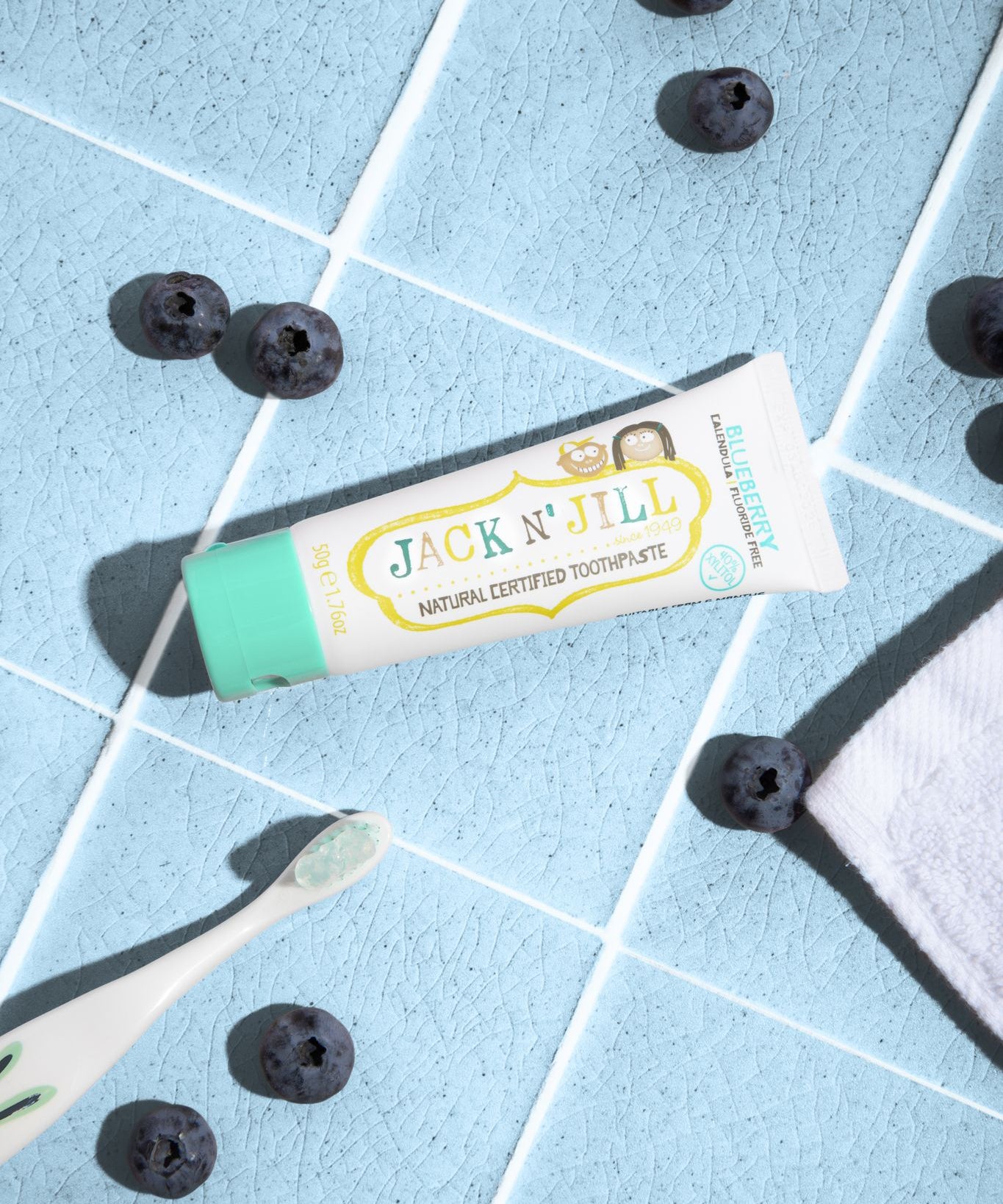 A 50g tube of Jack N' Jill blueberry flavoured fluoride free kid's toothpaste placed on a blue tiled surface next to a Jack N' Jill toothbrush surrounded by blueberries.