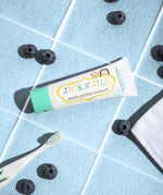 A 50g tube of Jack N' Jill blueberry flavoured fluoride free kid's toothpaste placed on a blue tiled surface next to a Jack N' Jill toothbrush surrounded by blueberries.