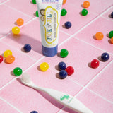 The Jack N Jill kids natural toothpaste in a bubblegum flavour, on pale pink tiles surrounded by colourful pieces of bubblegum and next to a kids toothbrush.