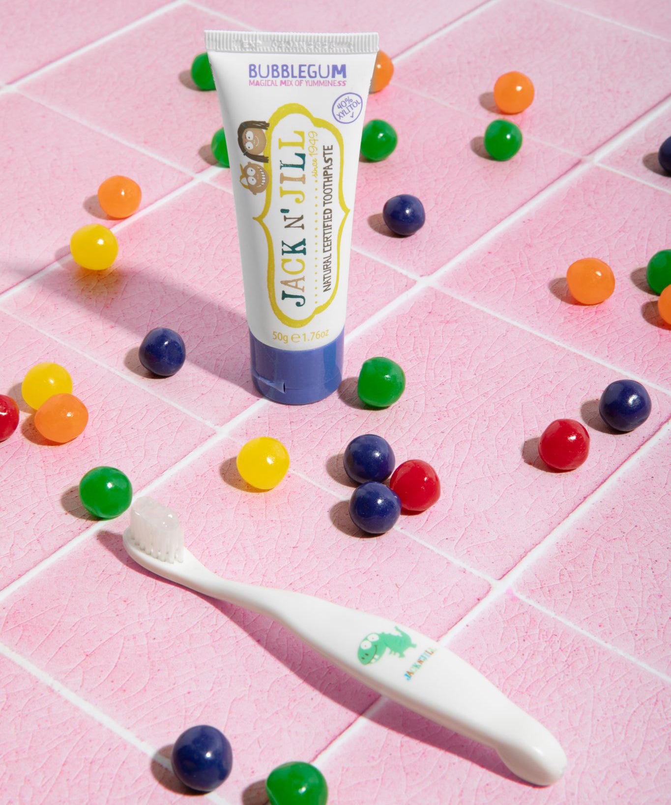 The Jack N Jill kids natural toothpaste in a bubblegum flavour, on pale pink tiles surrounded by colourful pieces of bubblegum and next to a kids toothbrush.