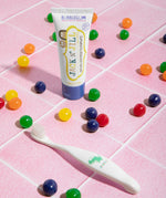 The Jack N Jill kids natural toothpaste in a bubblegum flavour, on pale pink tiles surrounded by colourful pieces of bubblegum and next to a kids toothbrush.