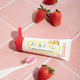 A 50g tube of Jack N' Jill strawberry flavoured fluoride free kid's toothpaste placed on a pink tiled surface next to a Jack N' Jill toothbrush and strawberries.