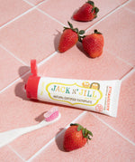 A 50g tube of Jack N' Jill strawberry flavoured fluoride free kid's toothpaste placed on a pink tiled surface next to a Jack N' Jill toothbrush and strawberries.