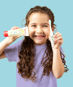 A child grinning and holding up a  50g tube of Jack N' Jill strawberry flavoured fluoride free kid's toothpaste next to a toothbrush.