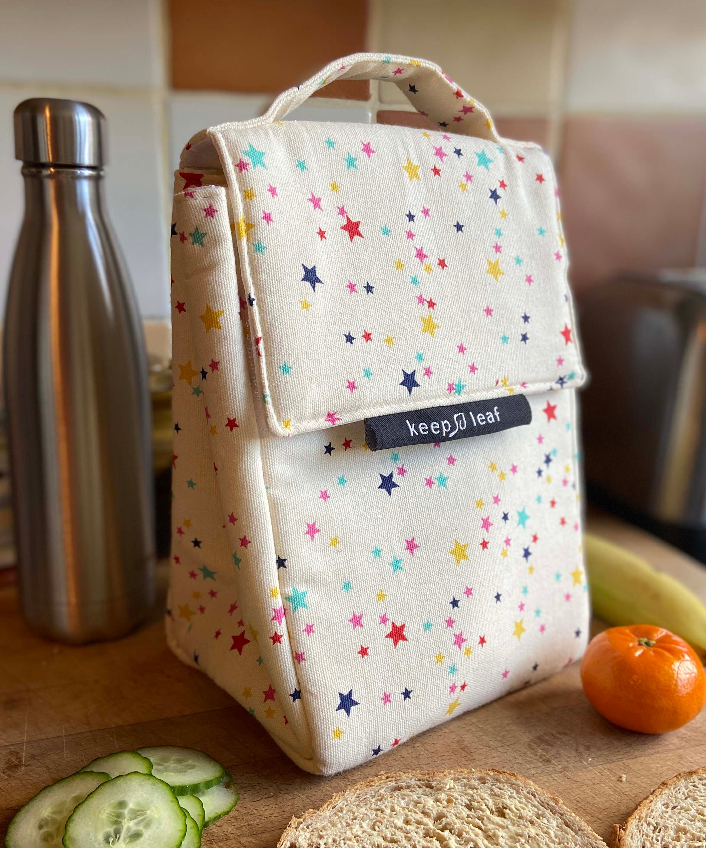 Keep Leaf insulated lunchbag in stars design in the kitchen on a wooden surface surrounded by food and a drinking bottle for size reference.