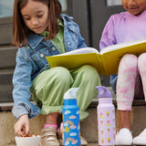 Two children sitting on a step outside, they have two Kid Kanteen 12oz Insulated Flip Seal Sport Cap Bottles in front of them. One bottle has a rainbow print, the other has an ice cream print.
