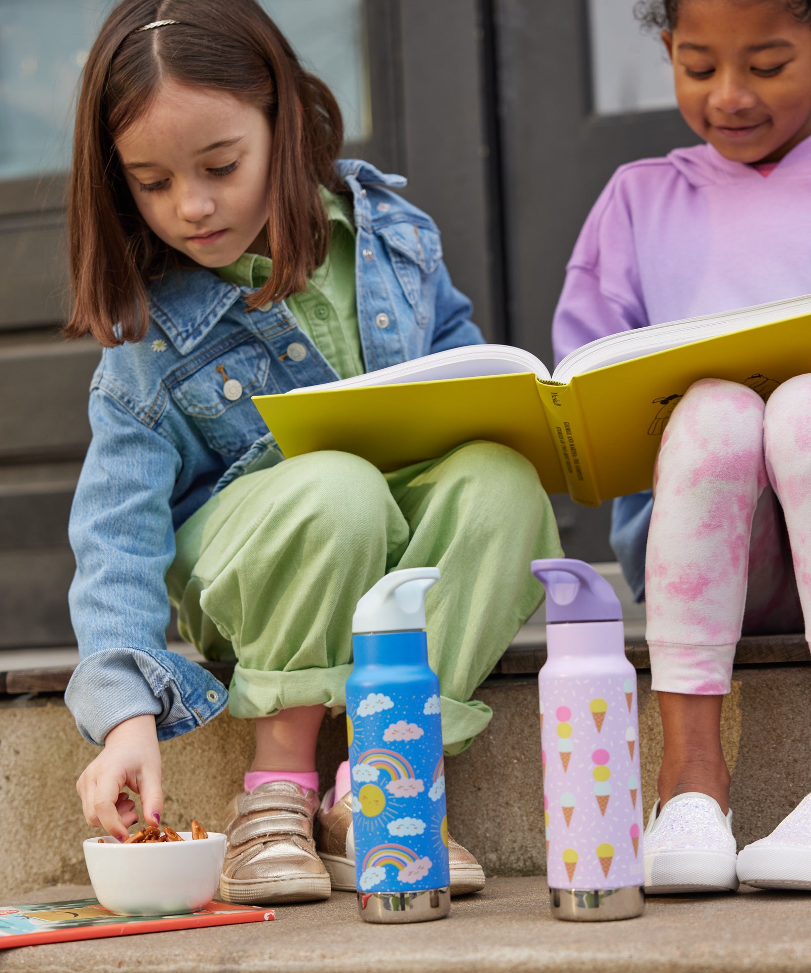 Two children sitting on a step outside, they have two Kid Kanteen 12oz Insulated Flip Seal Sport Cap Bottles in front of them. One bottle has a rainbow print, the other has an ice cream print.
