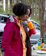 A woman standing outside in woodland and and drinking from marigold 16oz stainless steel reusable klean kanteen insulated rise bottle showing the colour way design detail with the Brittany Klean Kanteen bottle in the background.
