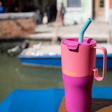 A close up of the Klean Kanteen 26oz rise insulated tumbler on a wooden surface near a river showing the pink colour way design and the flip lid detail with turquoise straw and handle detail