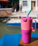 A close up of the Klean Kanteen 26oz rise insulated tumbler on a wooden surface near a river showing the pink colour way design and the flip lid detail with turquoise straw and handle detail