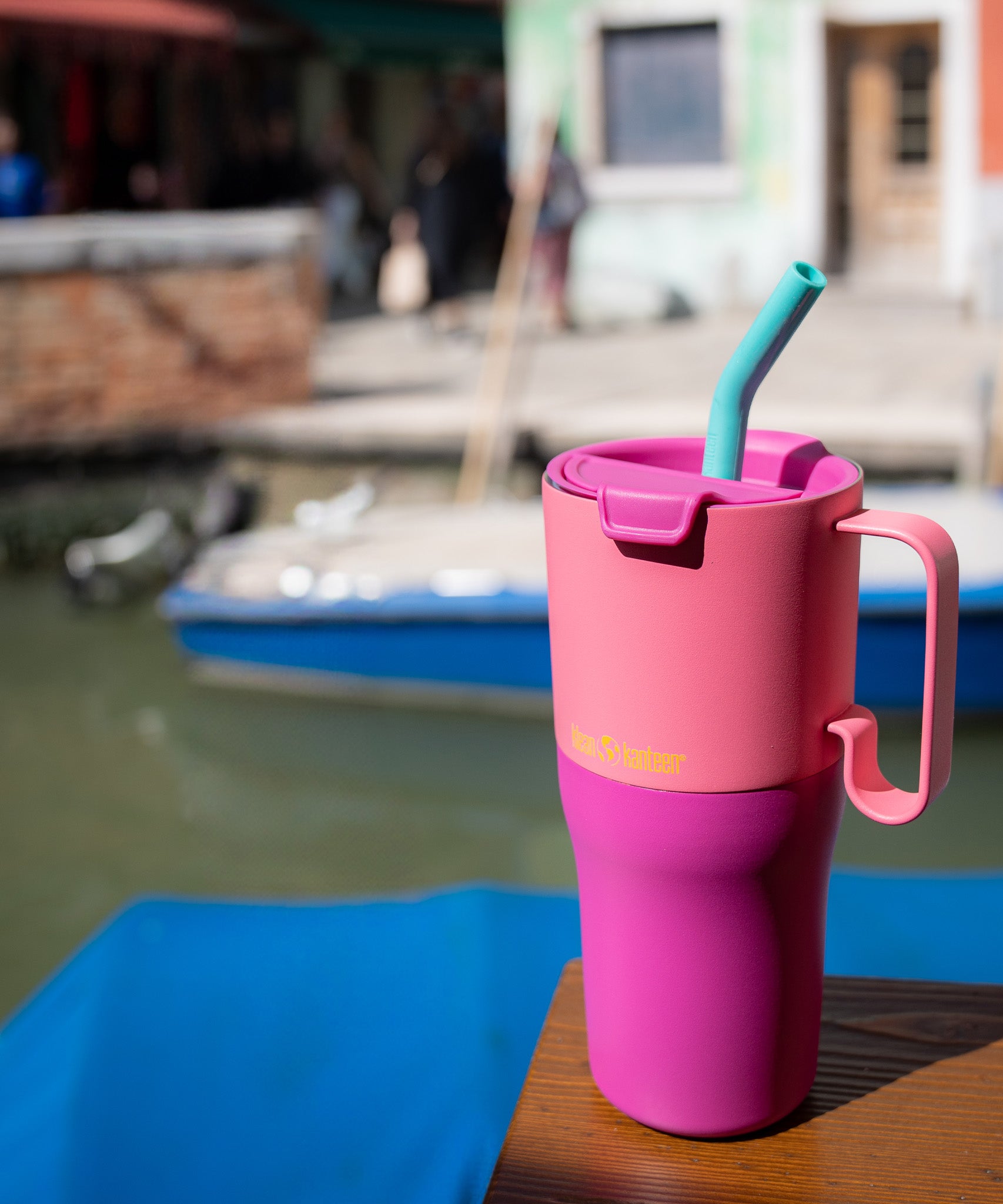 A close up of the Klean Kanteen 26oz rise insulated tumbler on a wooden surface near a river showing the pink colour way design and the flip lid detail with turquoise straw and handle detail