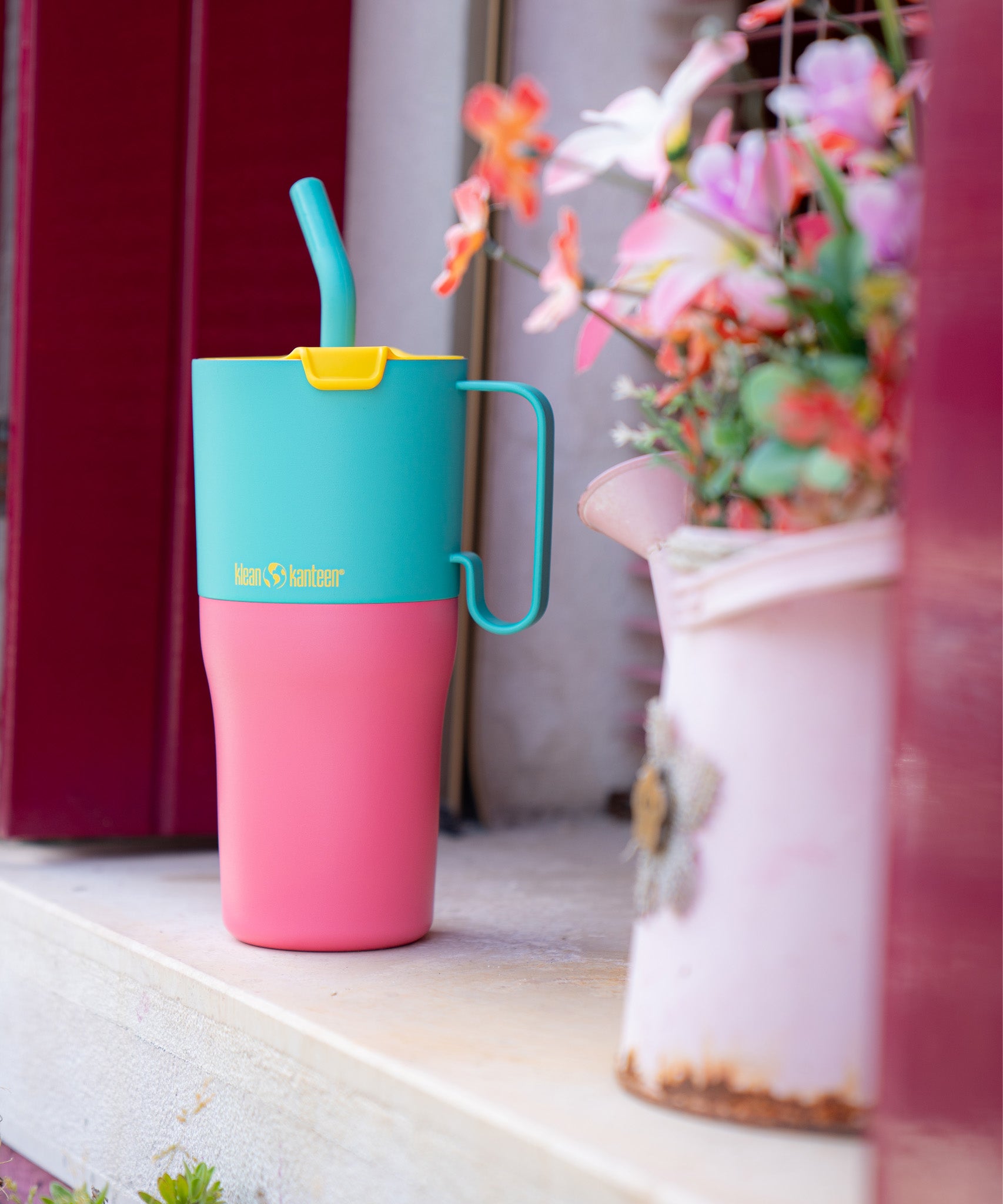 A close up of the Klean Kanteen 26oz turquoise stainless steel insulated rise tumbler on a windowsill showing the pink and turquoise colour way with a yellow flip cap. Features a turquoise handle on the side with a matching straw