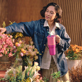 A women standing in a garden center surrounded by pretty flowers and holding the Klean Kanteen 36oz pink lemonade rise insulated tumbler by the handle on the side