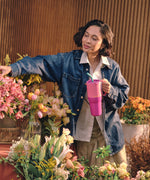 A women standing in a garden center surrounded by pretty flowers and holding the Klean Kanteen 36oz pink lemonade rise insulated tumbler by the handle on the side