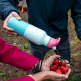 A adult holding the bay kaleidoscope 16oz Klean Kanteen insulated rise bottle by the bottom and pouring water into a handful of strawberries.