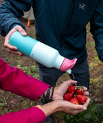 A adult holding the bay kaleidoscope 16oz Klean Kanteen insulated rise bottle by the bottom and pouring water into a handful of strawberries.