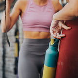 A woman standing in a gym environment and holding the Klean Kanteen 16oz Brittany kaleidoscope stainless steel reusable insulated rise bottle