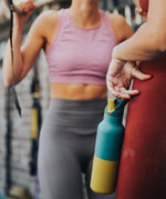 A woman standing in a gym environment and holding the Klean Kanteen 16oz Brittany kaleidoscope stainless steel reusable insulated rise bottle
