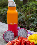 A close up of the marigold kaleidoscope 16oz Klean Kanteen reusable stainless steel rise bottle showing the orange and red colourway with white sports cap detail  on top of a wooden surface next to a bunch of fruit