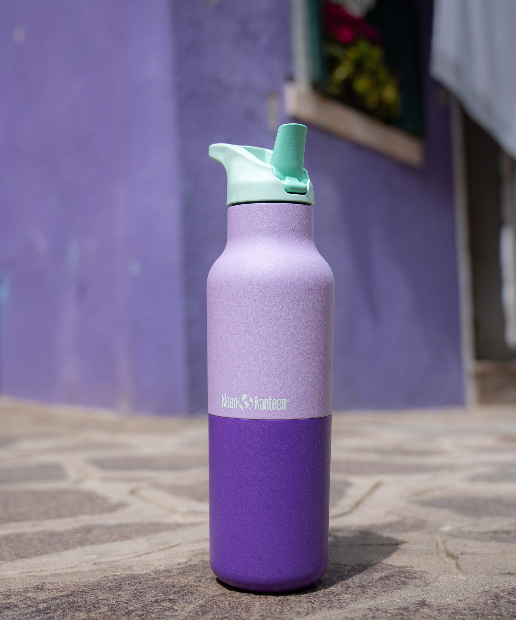 A close up of the orchid kaleidoscope 16oz klean kanteen insulated stainless steel rise bottle on a stone path showing the purple colour way with turquoise flilp sports cap detail