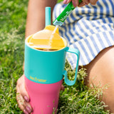 A close up of the Klean Kanteen 26oz turquoise stainless steel insulated rise tumbler on grass showing the yellow flip cap detail and the pink and turquoise colour way design