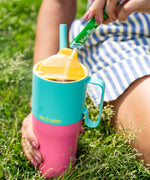 A close up of the Klean Kanteen 26oz turquoise stainless steel insulated rise tumbler on grass showing the yellow flip cap detail and the pink and turquoise colour way design