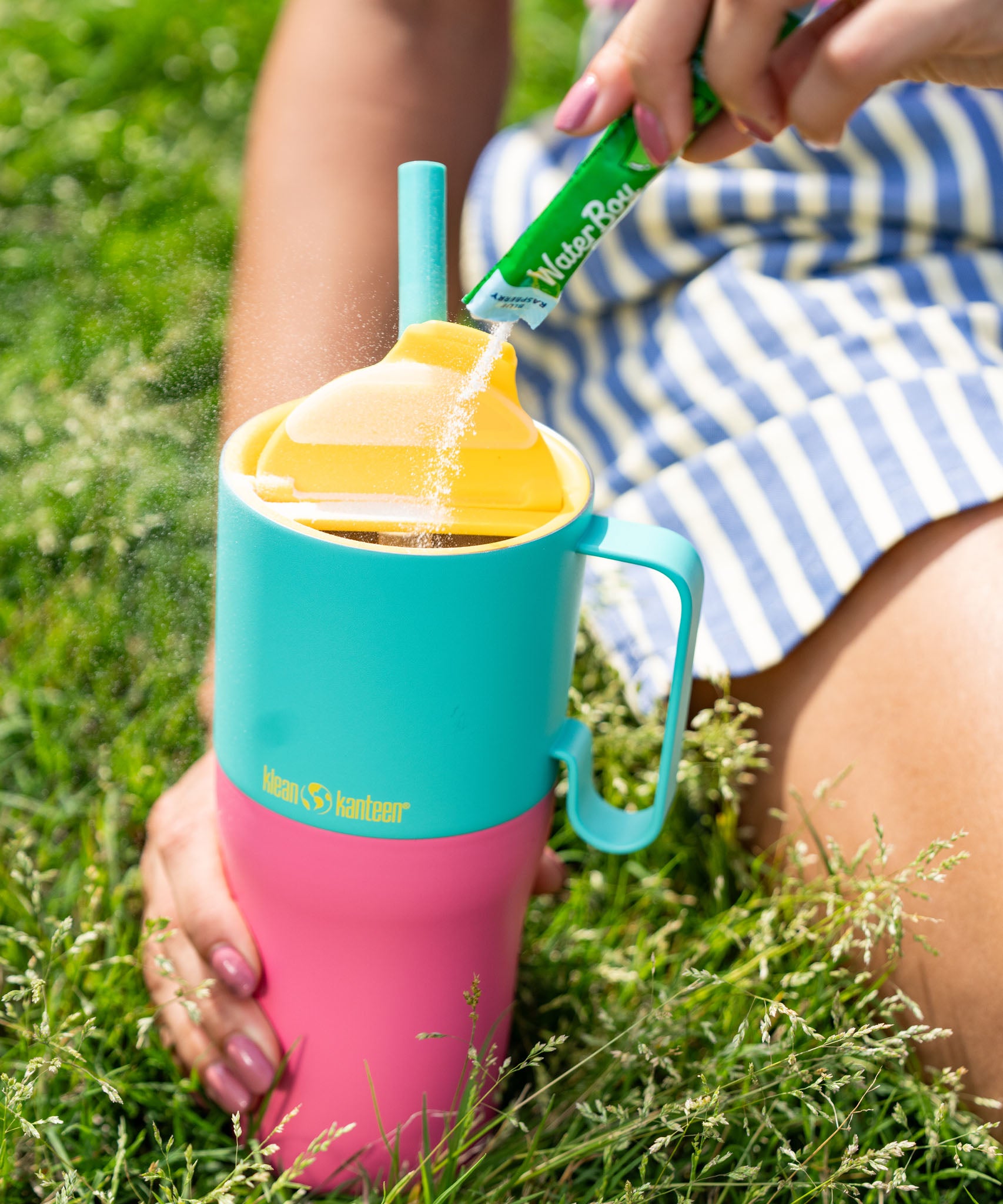 A close up of the Klean Kanteen 26oz turquoise stainless steel insulated rise tumbler on grass showing the yellow flip cap detail and the pink and turquoise colour way design