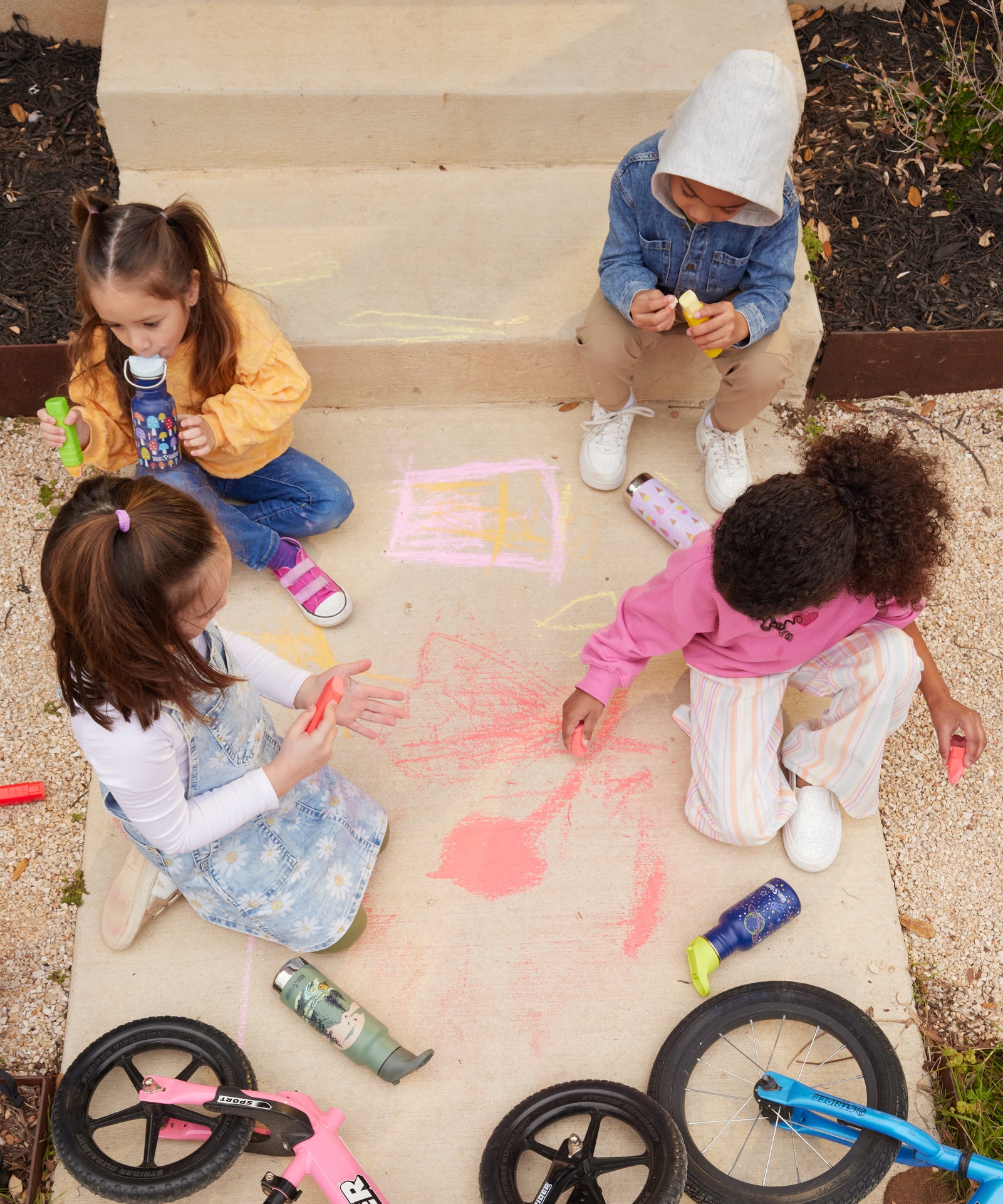 4 children sitting on a concreate path drawing with chalks, four different Kid Kanteen can be seen. One child is drinking out of a mushroom print sippy cap Kid Kanteen. There are 3 other Kid Kanteen on the ground next to the children, an insulated Ice Cream print bottle, an insulated Mountain Range bottle and a Milky Way print bottle.