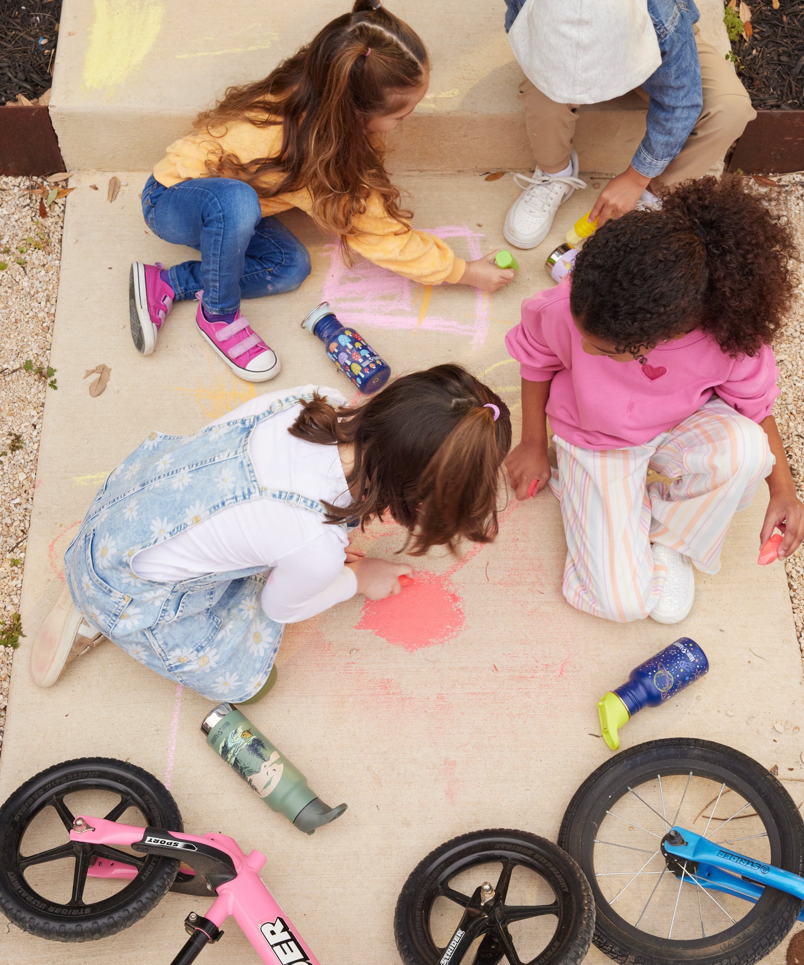 Children sitting on a concrete path drawing on the path with chalks. There are Kid Kanteen reusable water bottles on the ground next to the children.