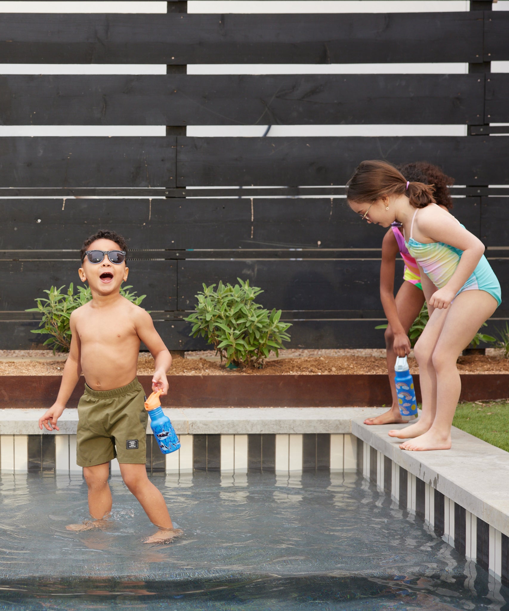Two children in a splash pool, one is holding the Sharks in Shades Kid Kanteen 12oz Narrow Flip Seal Sport Cap Bottle the other has an insulated Rainbow print Kid Kanteen Bottle.