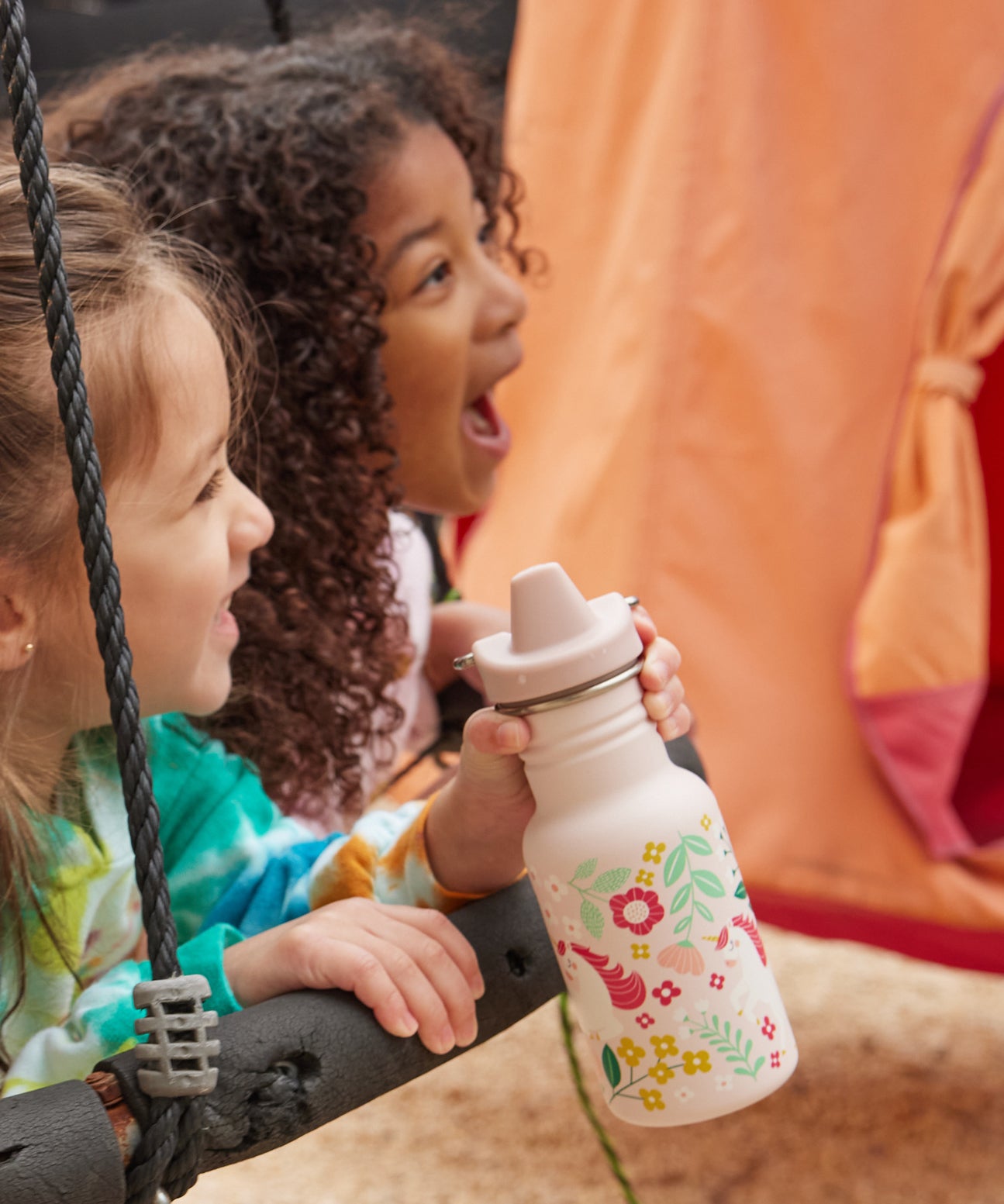 Two children on a swing, one child is holding a Kid Kanteen 12oz Narrow Sippy Bottle featuring a leaping unicorns print.