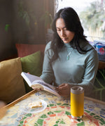 A person sitting at a moisaic covered table reading a book. There is A Klean Kanteen 12oz TKWide Cafe in the sunset yellow colour in front of them.