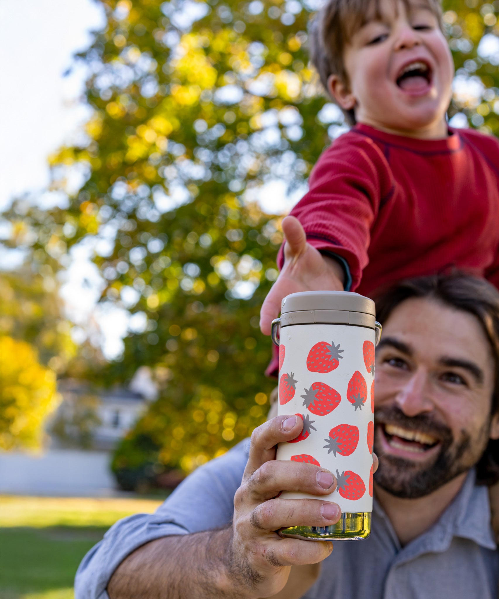 A child reaching for a Klean Kanteen 12oz Tofu Strawberries Print TKWide from an adult. The child is sitting on the adult's shoulders.