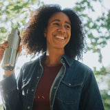 A woman standing under a tree and holding a opened klean Kanteen 16oz reflect stainless steel rise bottle