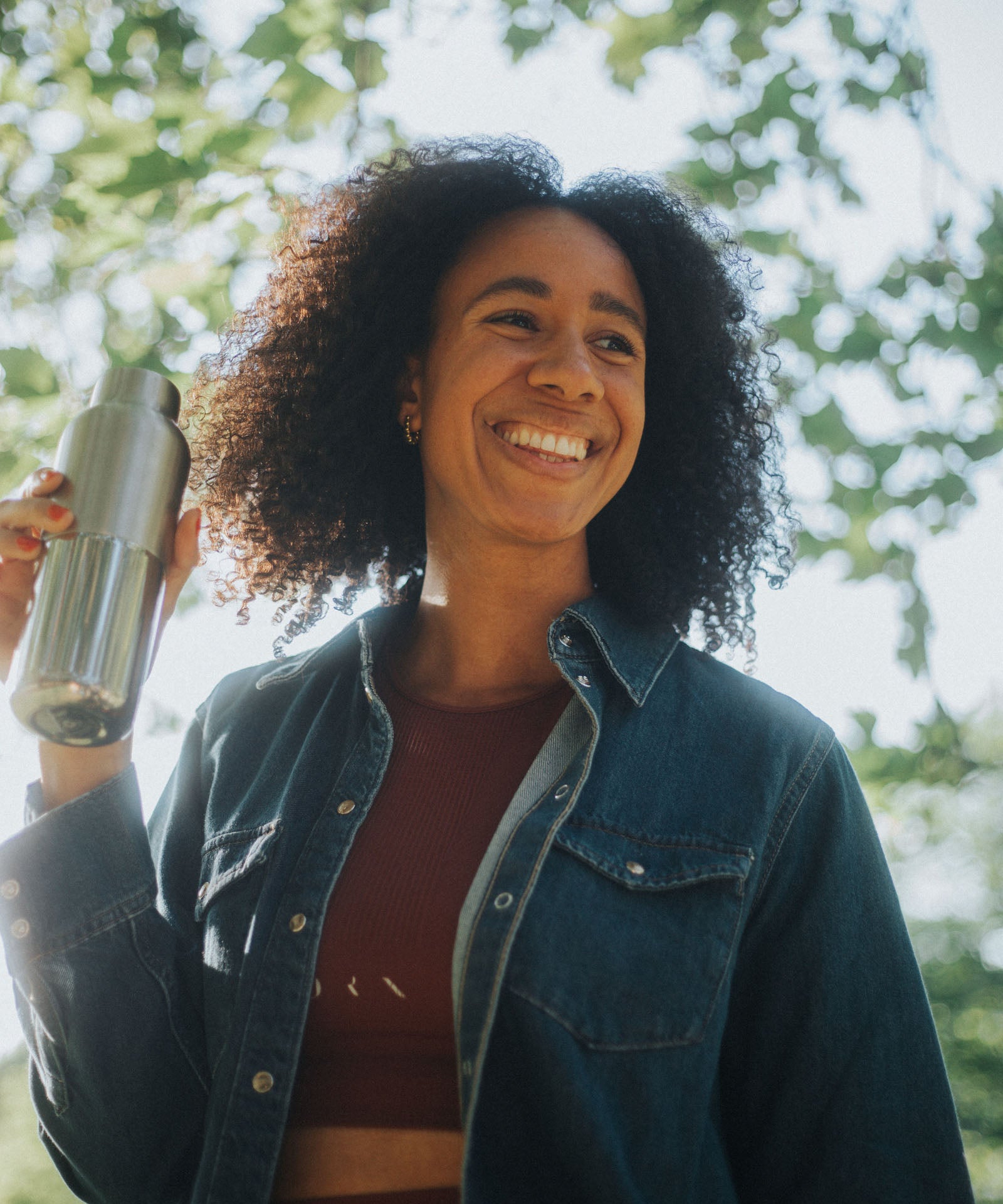 A woman standing under a tree and holding a opened klean Kanteen 16oz reflect stainless steel rise bottle