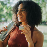A woman standing outside and holding a klean Kanteen 16oz reflect stainless steel rise bottle with the bamboo loop cap in the other hand