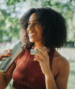 A woman standing outside and holding a klean Kanteen 16oz reflect stainless steel rise bottle with the bamboo loop cap in the other hand