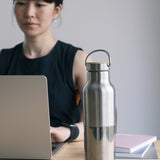 A woman sitting in a office environment  with the  klean Kanteen 16oz reflect stainless steel rise bottle on a wooden table showing the finish and loop cap detail