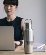 A woman sitting in a office environment  with the  klean Kanteen 16oz reflect stainless steel rise bottle on a wooden table showing the finish and loop cap detail