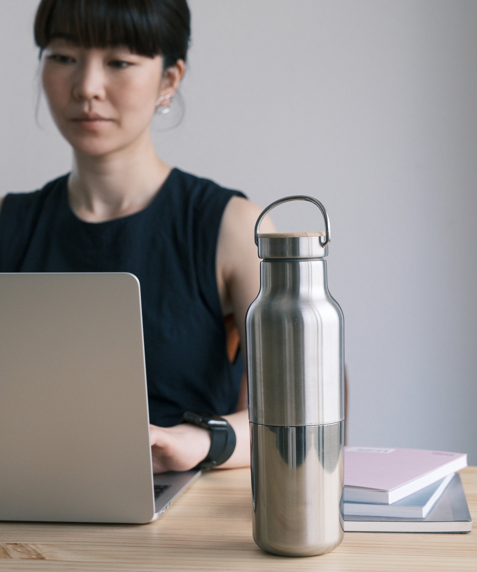 A woman sitting in a office environment  with the  klean Kanteen 16oz reflect stainless steel rise bottle on a wooden table showing the finish and loop cap detail