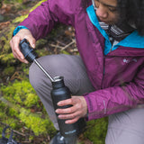 A person kneeling down holding a black coloured Klean Kanteen 18oz Classic Narrow Flip Seal Sports Cap Reusable Stainless Steel Water Bottle in their hand with the lid off