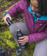 A person kneeling down holding a black coloured Klean Kanteen 18oz Classic Narrow Flip Seal Sports Cap Reusable Stainless Steel Water Bottle in their hand with the lid off