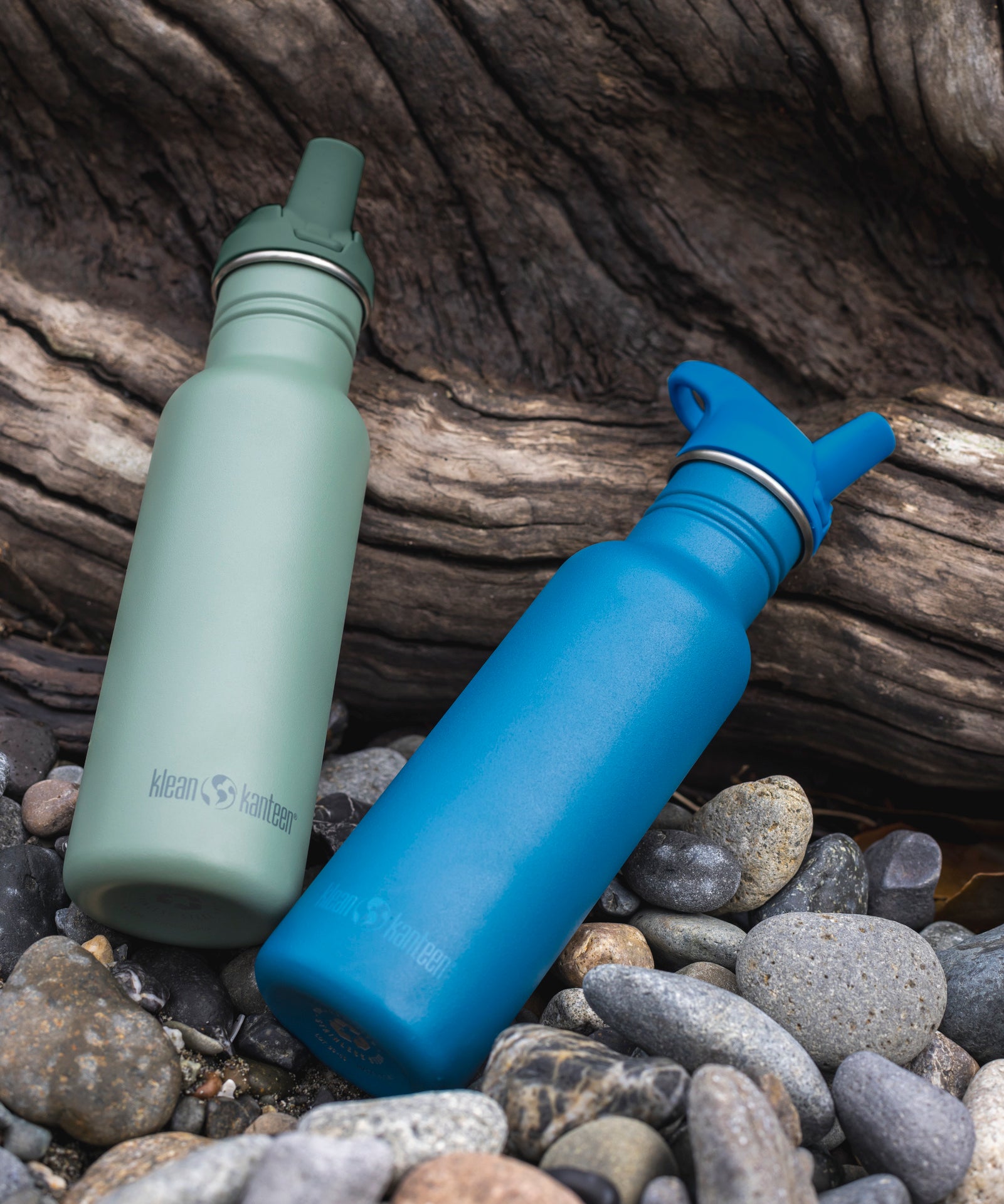 Two Klean Kanteen 18oz Classic Narrow Flip Seal Sports Cap Reusable Stainless Steel Water Bottle resting on stones and driftwood. One bottle is in the Sea Spray green colour and the other is the Corsair blue colour.