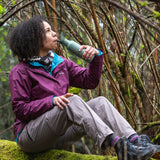 A person sitting on a moss covered surface drinking from a Sea Spray green coloured Klean Kanteen 18oz Classic Narrow Flip Seal Sports Cap Reusable Stainless Steel Water Bottle.