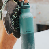 A climber holding the Klean Kanteen 20oz rise insulated Reusable Stainless Steel Water Bottle with loop cap in a Brittany blue colour.