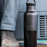 A close up of the black 20oz Klean Kanteen Reusable Stainless Steel Water Bottle next to a adult leg showing the size and coating detail