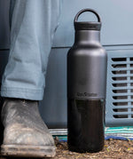 A close up of the black 20oz Klean Kanteen Reusable Stainless Steel Water Bottle next to a adult leg showing the size and coating detail