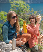 Two people sitting on rocks, one person is holding a Sunset yellow coloured Klean Kanteen 27oz Classic Narrow Flip Seal Sports Cap Reusable Stainless Steel Water Bottle there is another brushed steel bottle on the rock in front of them.