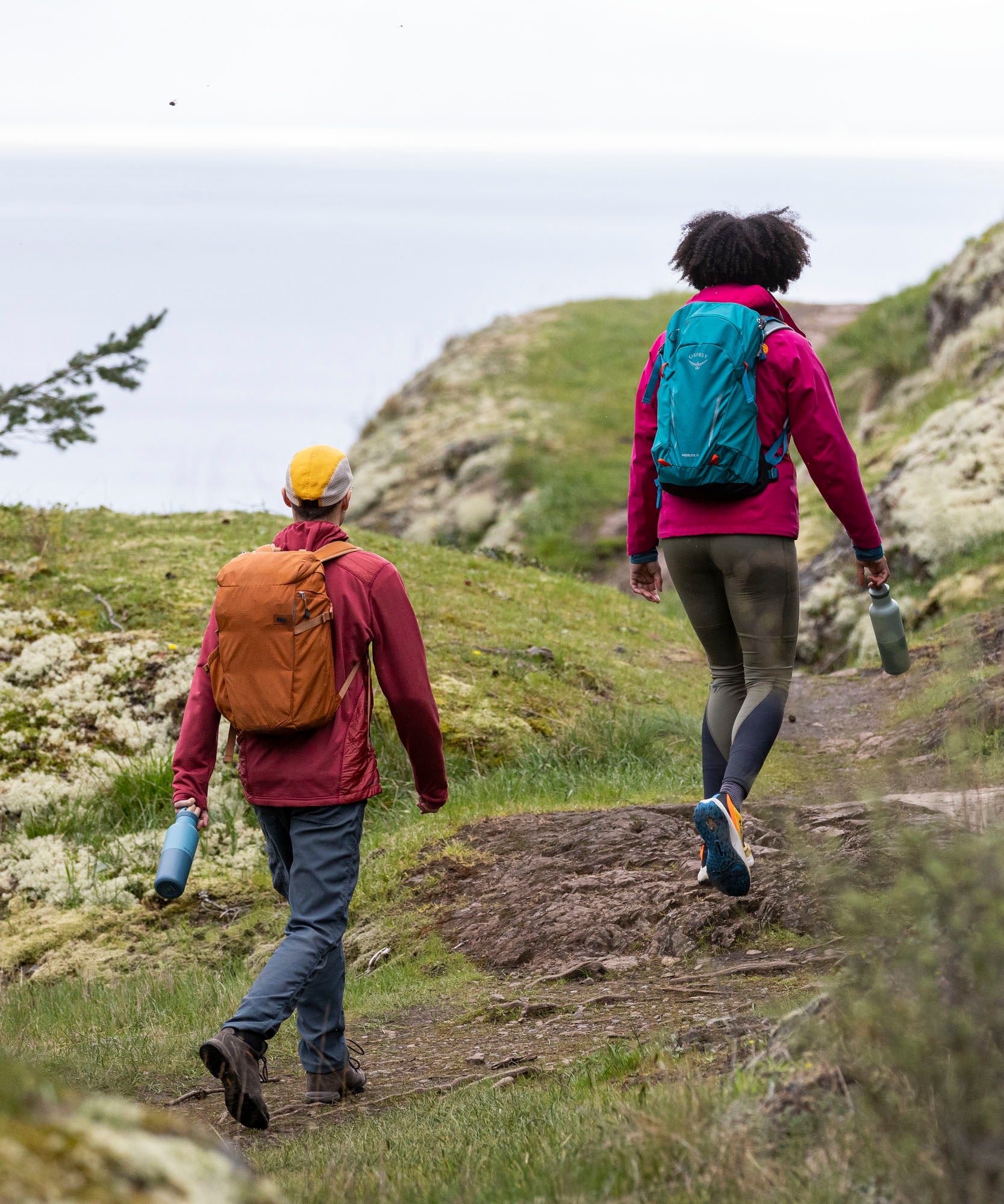 2 adult walkers outside walking on a cliff and holding the Brittany blue and iceberg stainless reusable water Klean Kanteen 20oz bottles