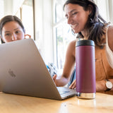 Close up of a 16oz Klean Kanteen reusable metal bottle in front of two girls using a laptop on a wooden table
